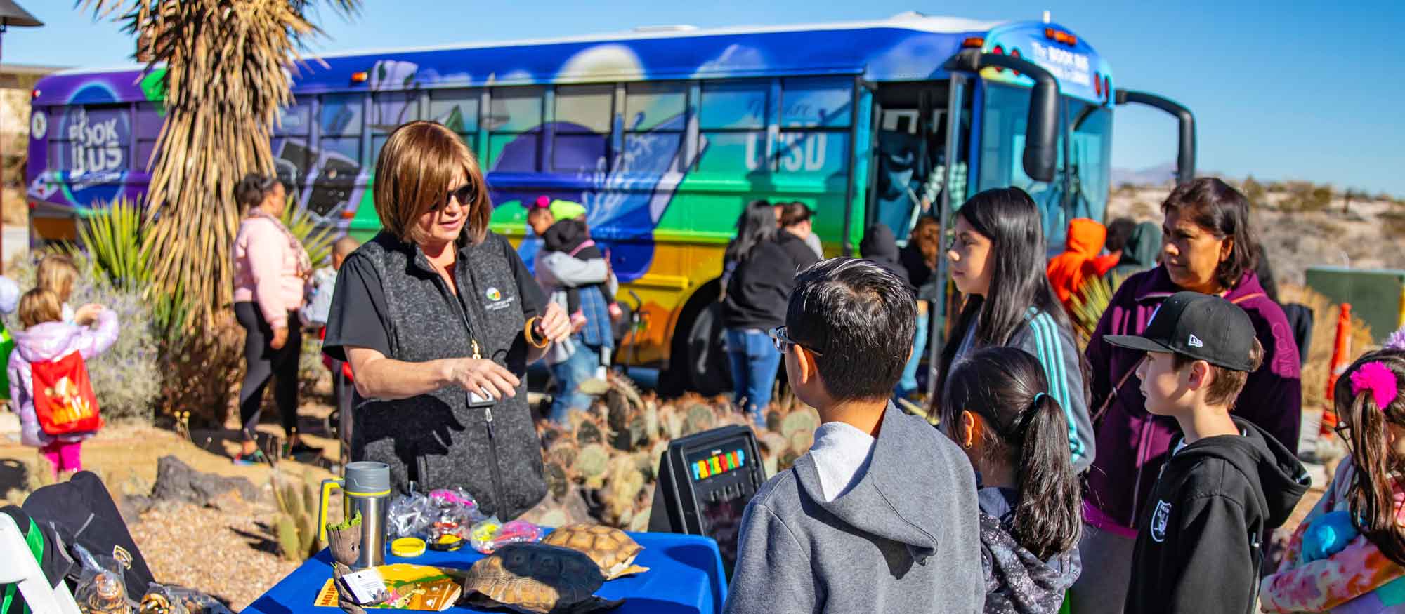 A person is interacting with children at an outdoor educational event with a table displaying various items. In the background, a bus with vibrant colors and signs is visible.