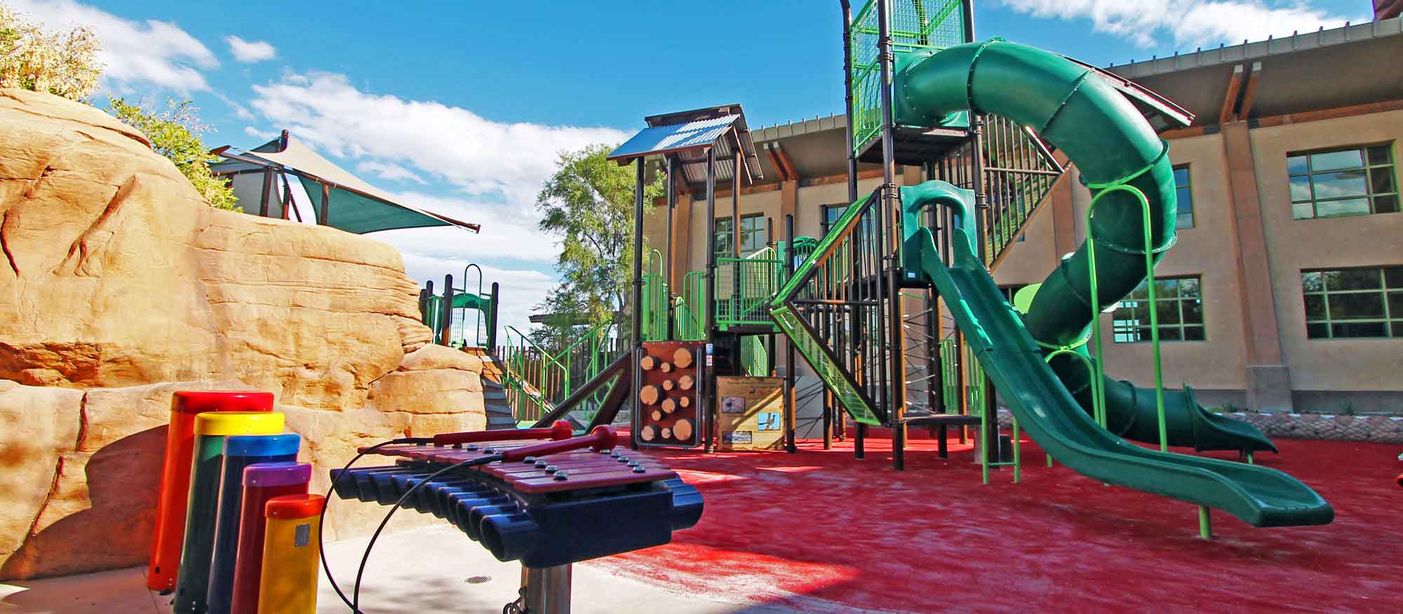 An outdoor playground featuring a tall green slide, climbing structures, and musical play equipment on a red safety surface, set against a backdrop of a clear sky.