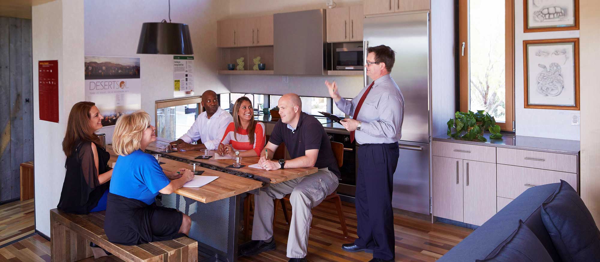 Five adults are engaged in a meeting around a wooden table in a stylish, modern kitchen area. There's contemporary artwork on the walls and a comfortable seating area to the side.