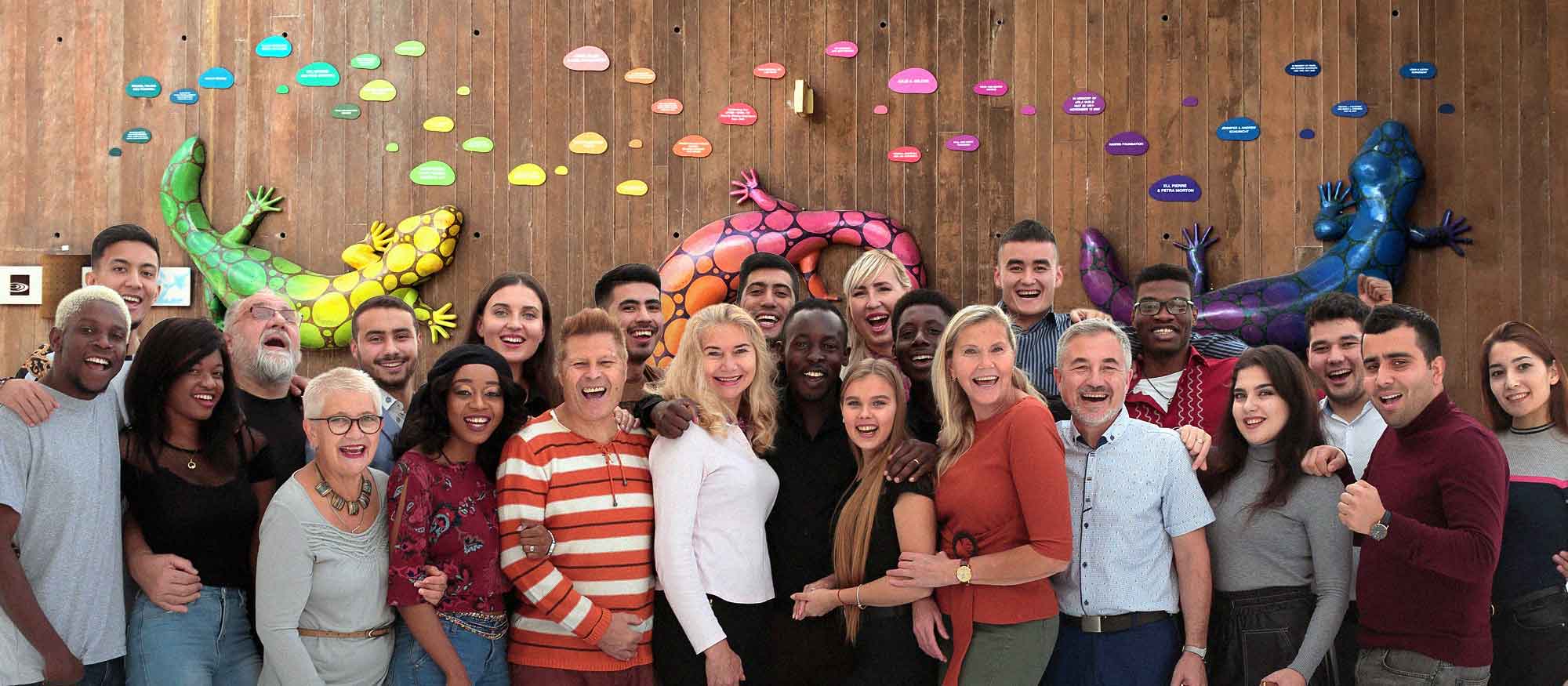 Group of diverse individuals smiling and posing together in front of a wooden donor recognition wall decorated with colorful Gila monster lizard sculptures.