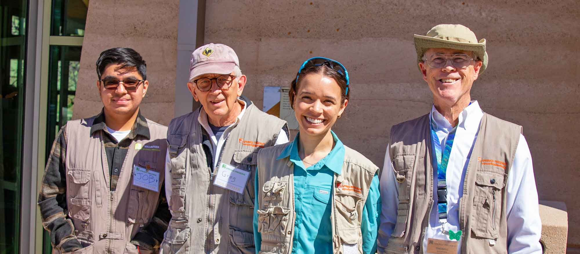 Four volunteers wearing vests and name tags stand smiling outside a building.