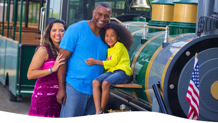 A family of three smiling near a vintage-style trackless train.