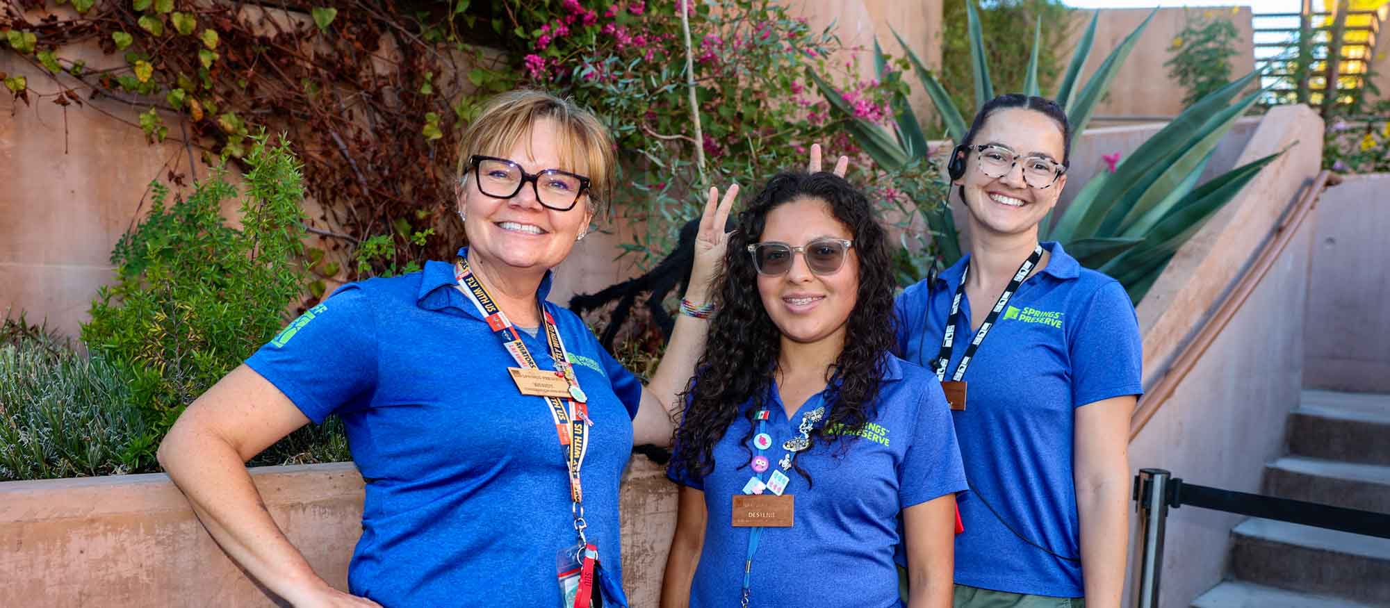 Three women wearing blue shirts and name tags are smiling in an outdoor setting with plants in the background.
