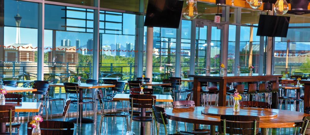 Interior of a bright, spacious restaurant with tables set for dining, overlooking a cityscape visible through large windows.