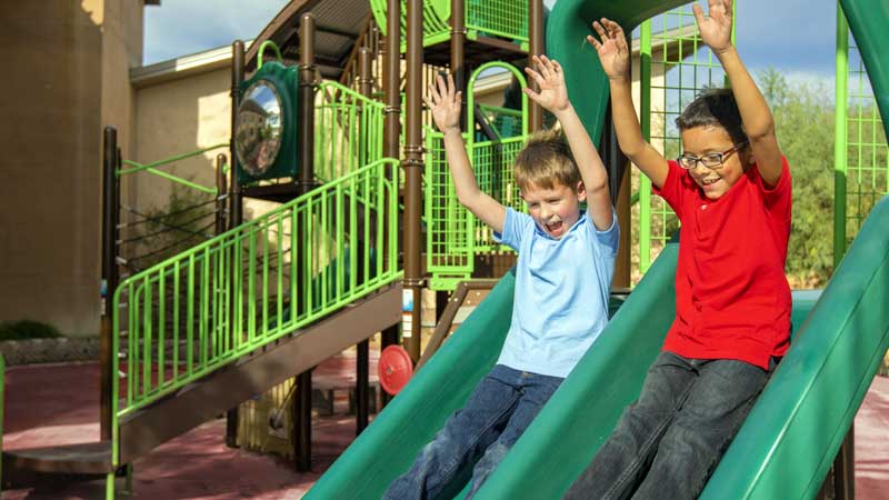 Two children joyfully sliding down a green slide at a playground.