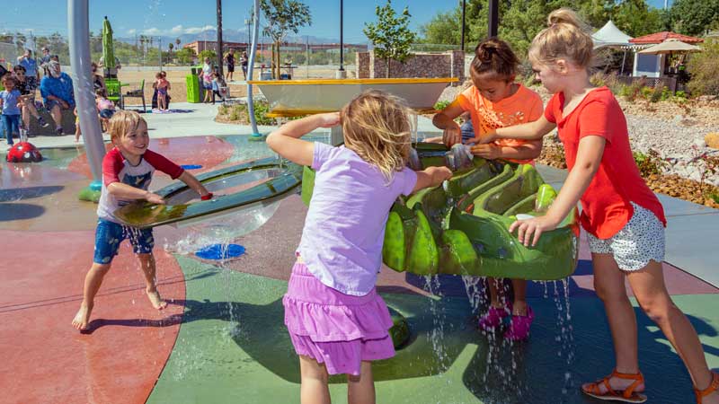 Children playing with water features at a colorful splash pad park on a sunny day.