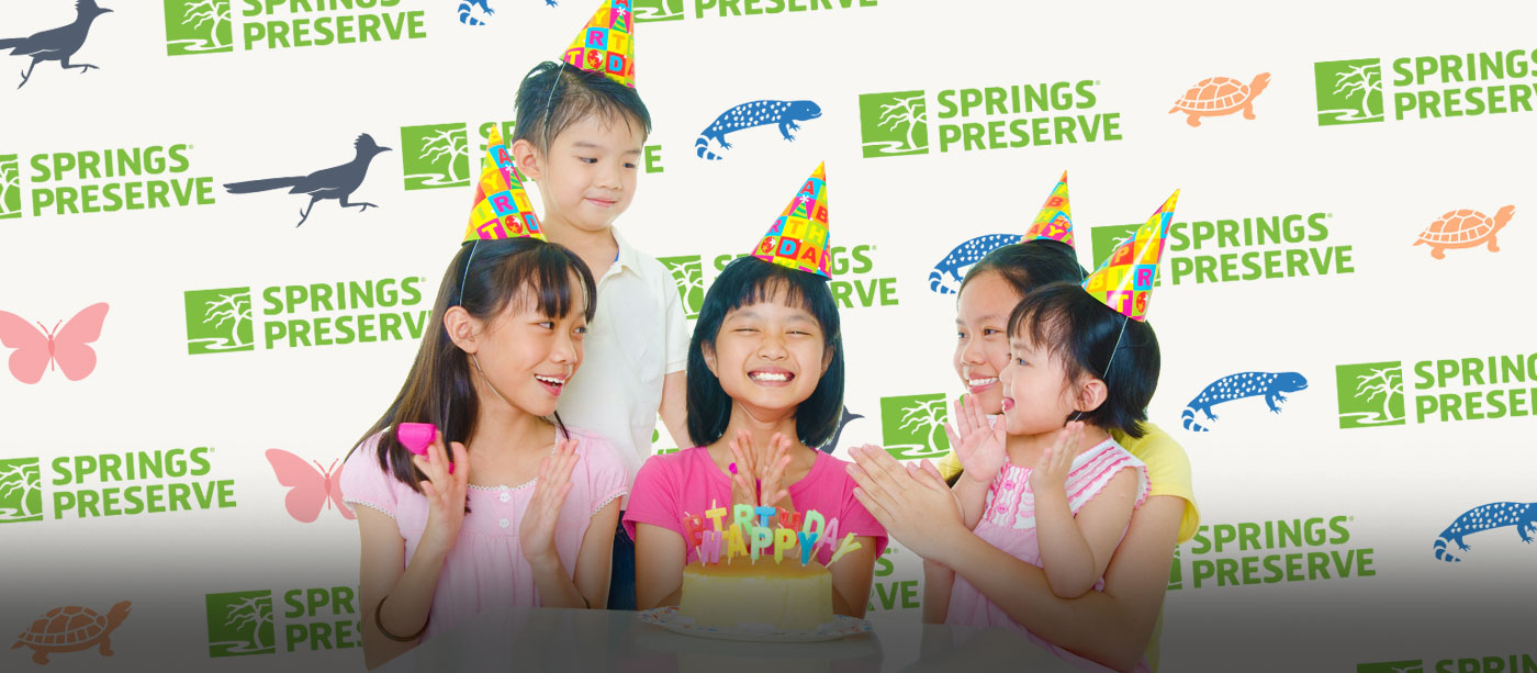 Birthday event at the Las Vegas Springs Preserve shows 5 children wearing festive hats in front of birthday cake smiling and clapping