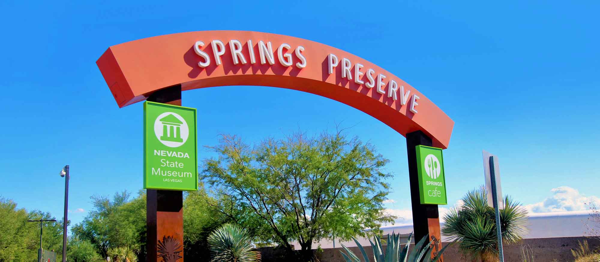 Entrance archway to Springs Preserve with signs for Nevada State Museum on a sunny day, flanked by desert landscaping.