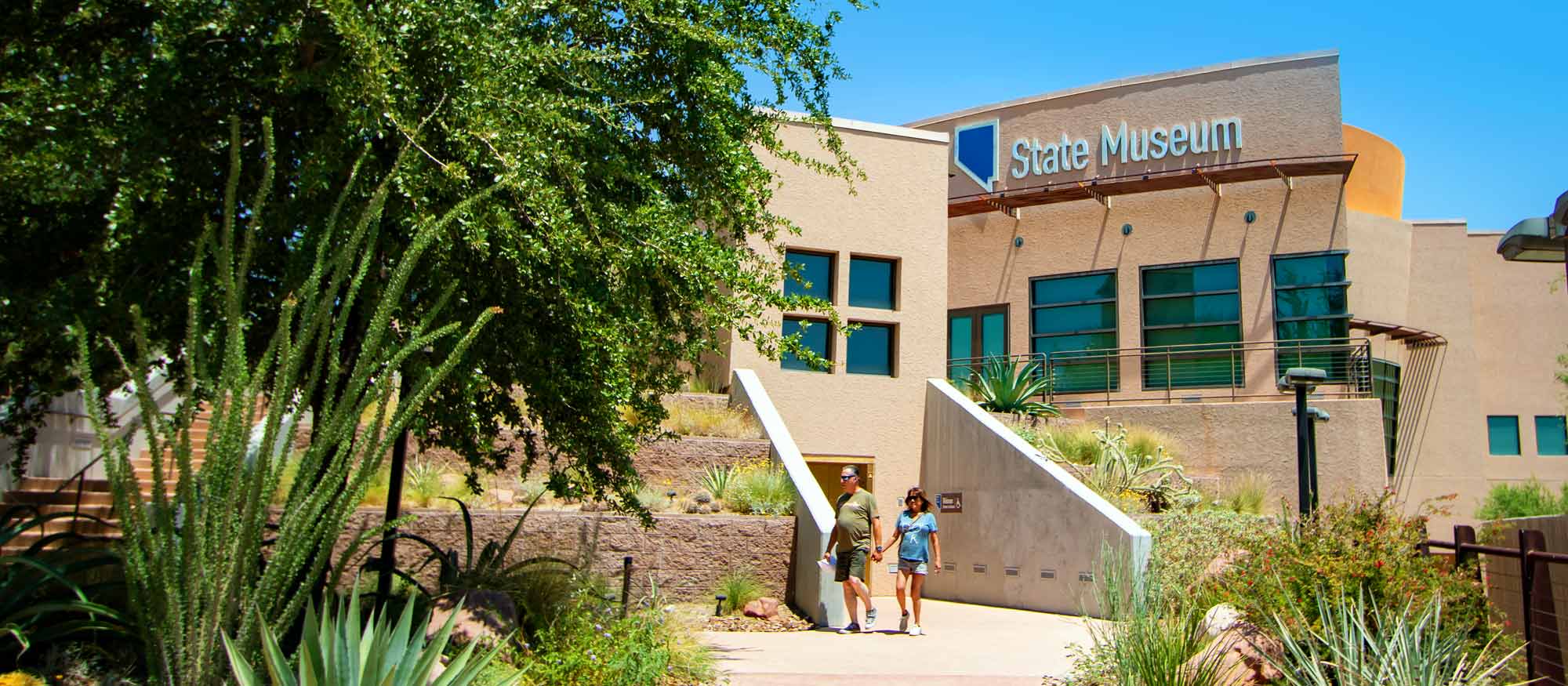 Exterior view of the Nevada State Museum on a sunny day, featuring desert landscaping with cacti and a clear sky. Two visitors are walking towards the entrance.