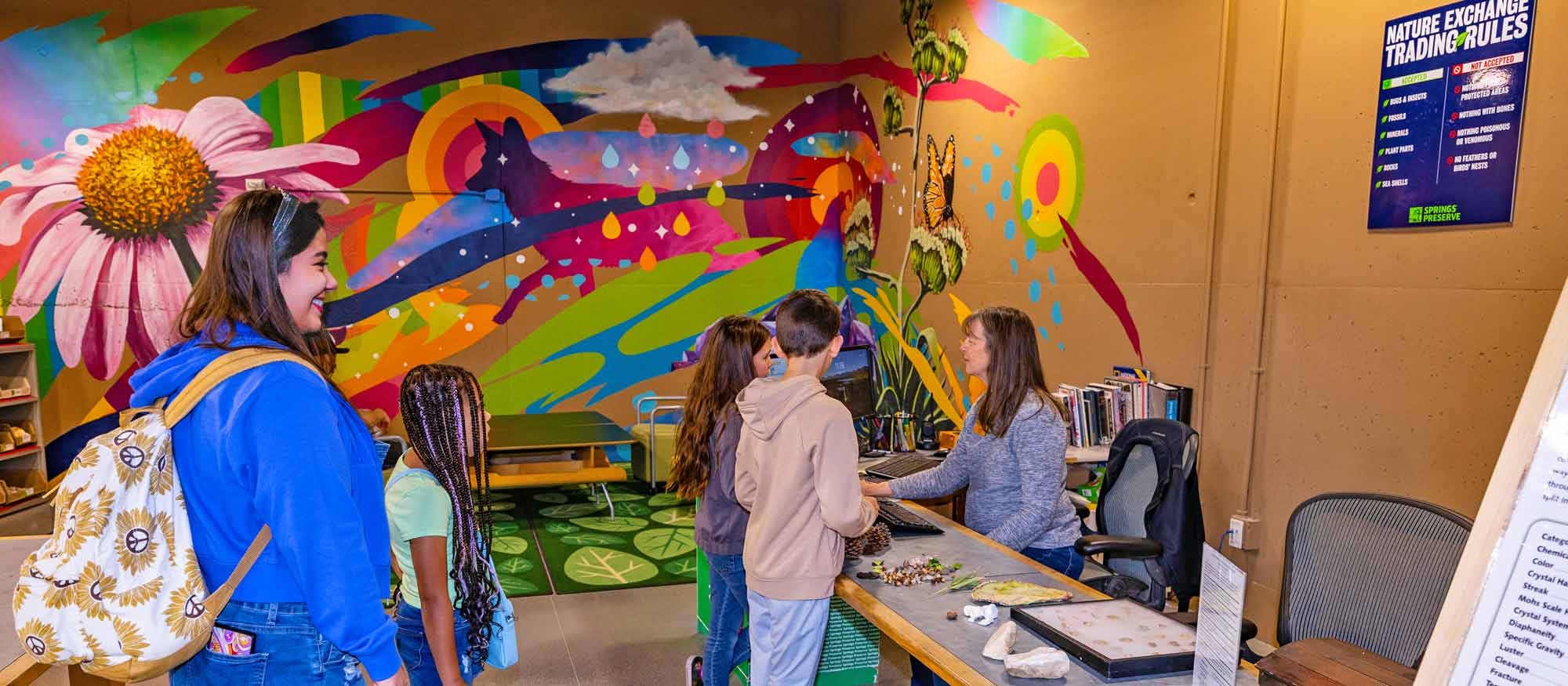 Visitors engaging with an interactive display at the Nature Exchange in a room with a vibrant, nature-themed mural on the wall.