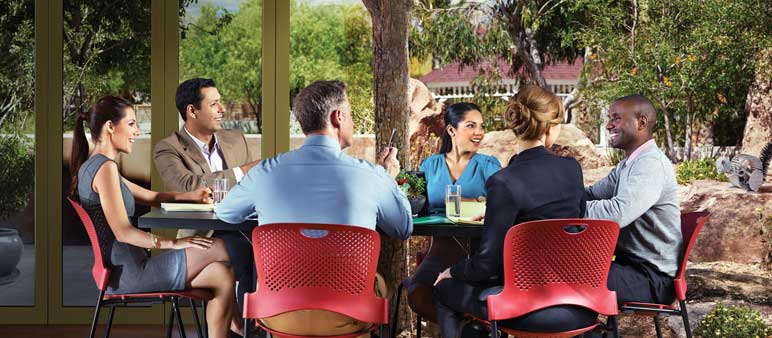 A group of six professionals is seated at tables outside, engaged in discussion near a tree-lined area. The setting includes open doors leading to an indoor meeting space.