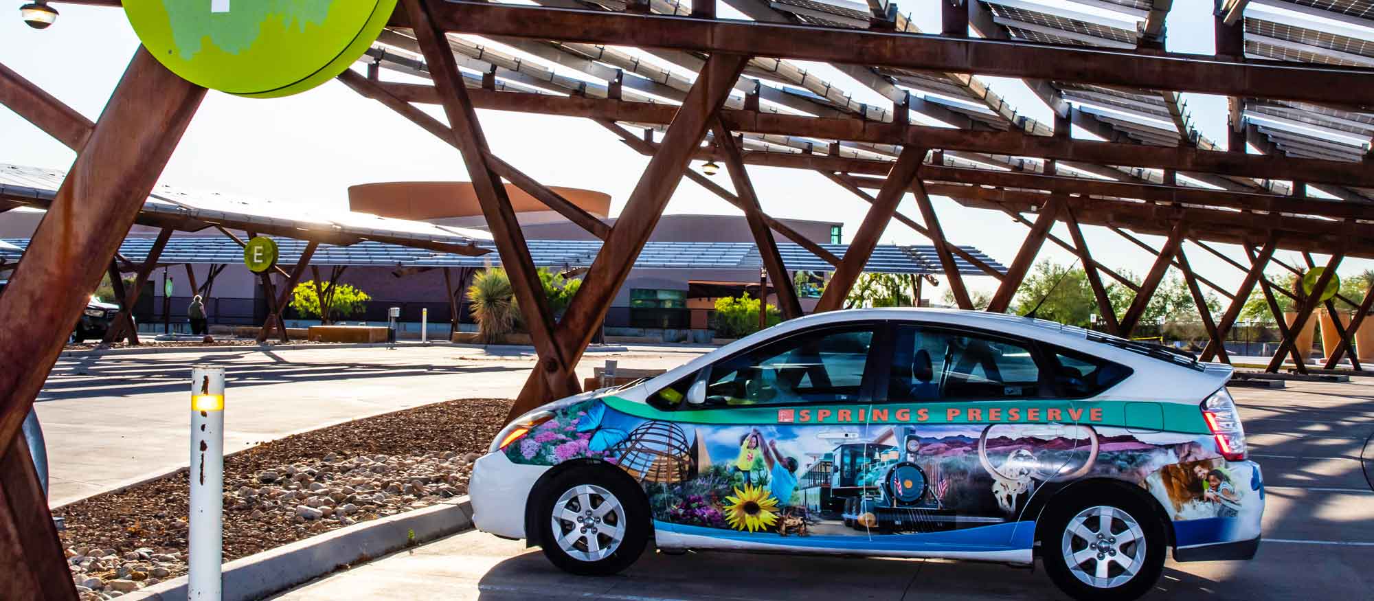 A colorful car with nature-themed graphics and Springs Preserve logo branding, parked under a large solar panel structure in a sunny outdoor setting.
