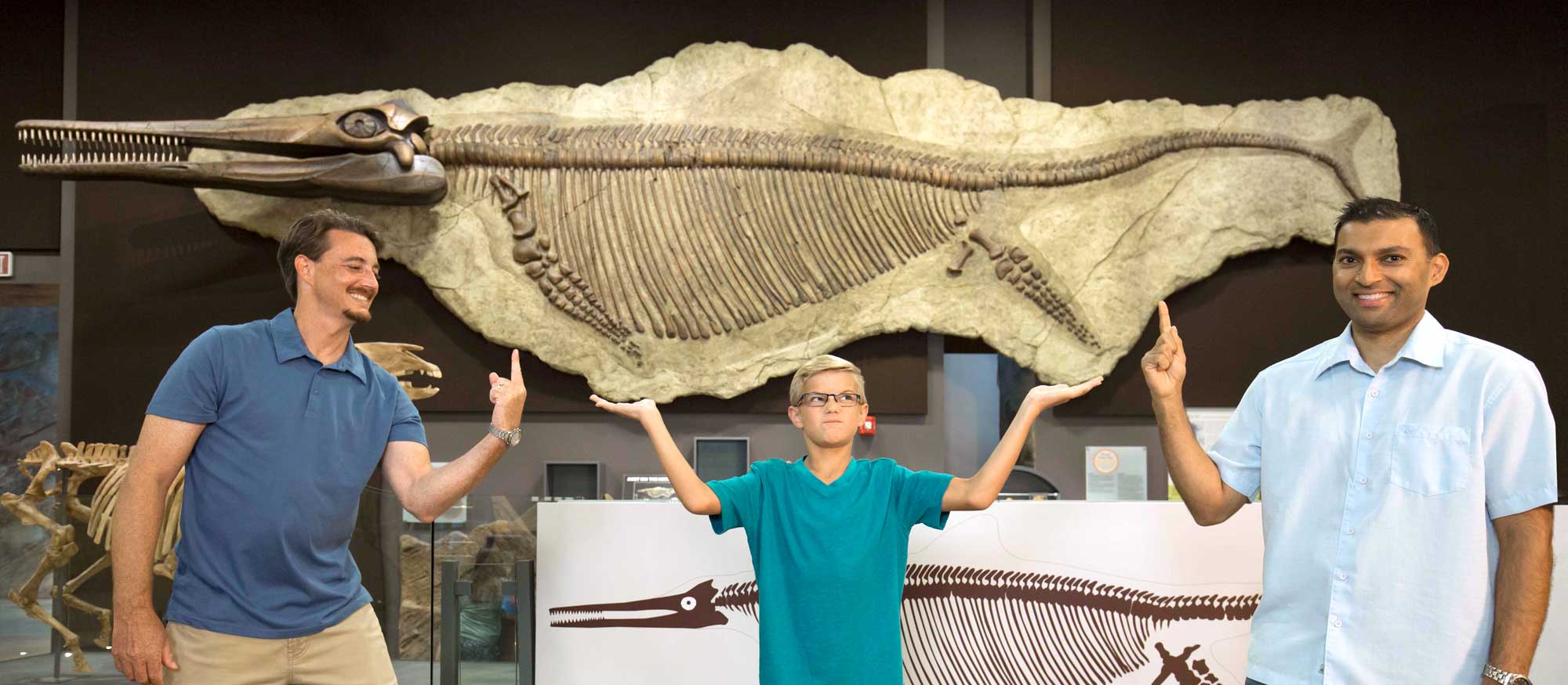 Three individuals pose in front of a large Ichthyosaur fossil displayed in a museum.