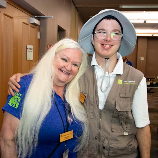 Two individuals, one wearing a blue shirt with a staff badge and the other in a beige volunteer vest and sun hat, smiling and posing together at an indoor event.