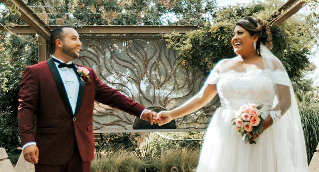 Two individuals in wedding attire holding hands and smiling at each other in an outdoor setting with lush greenery and a decorative backdrop. Photo courtesy Cascade and Canyon Photography.